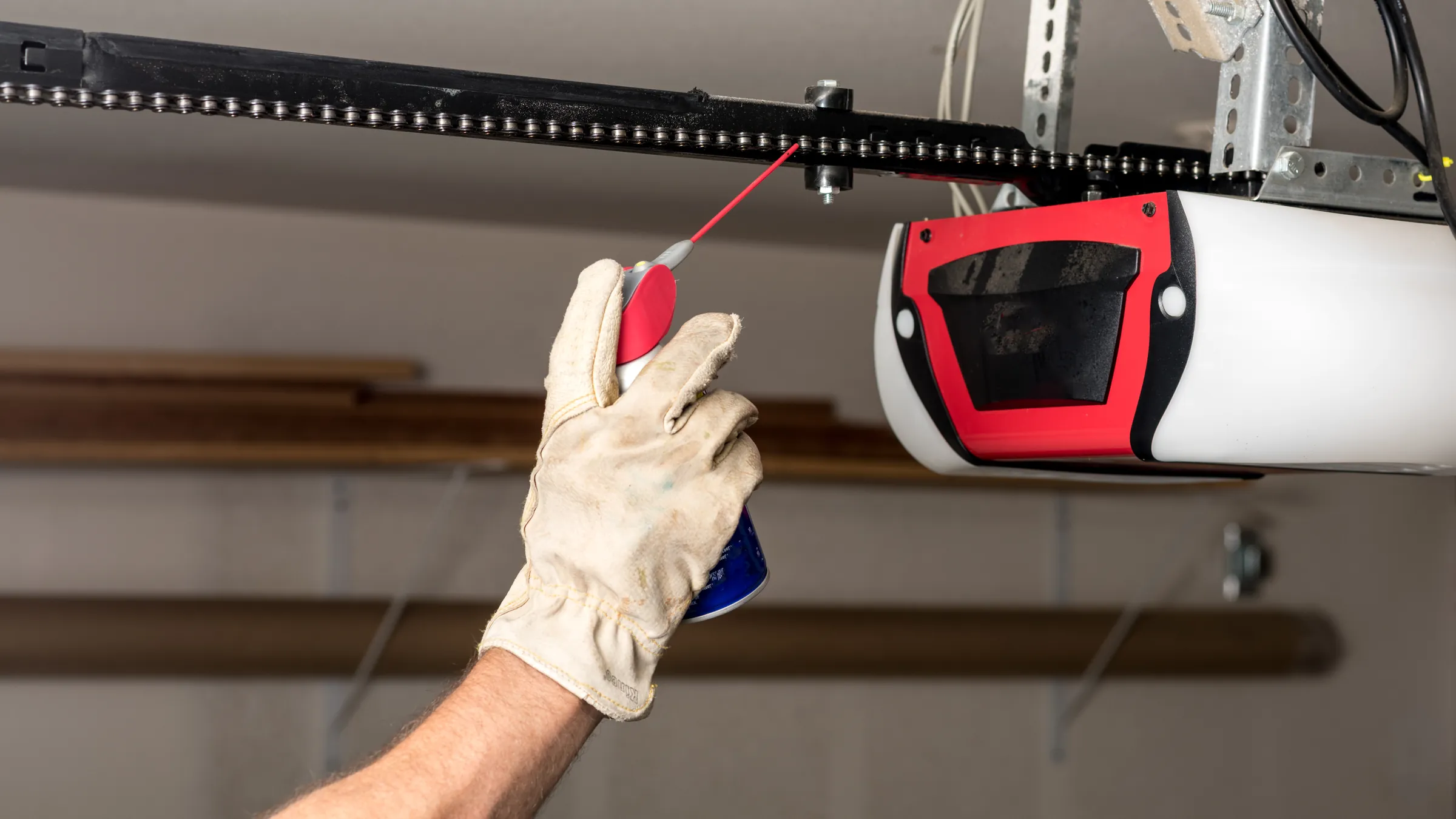 Gloved hand spraying lubricant onto a garage door opener's drive chain, beside the red and white opener unit