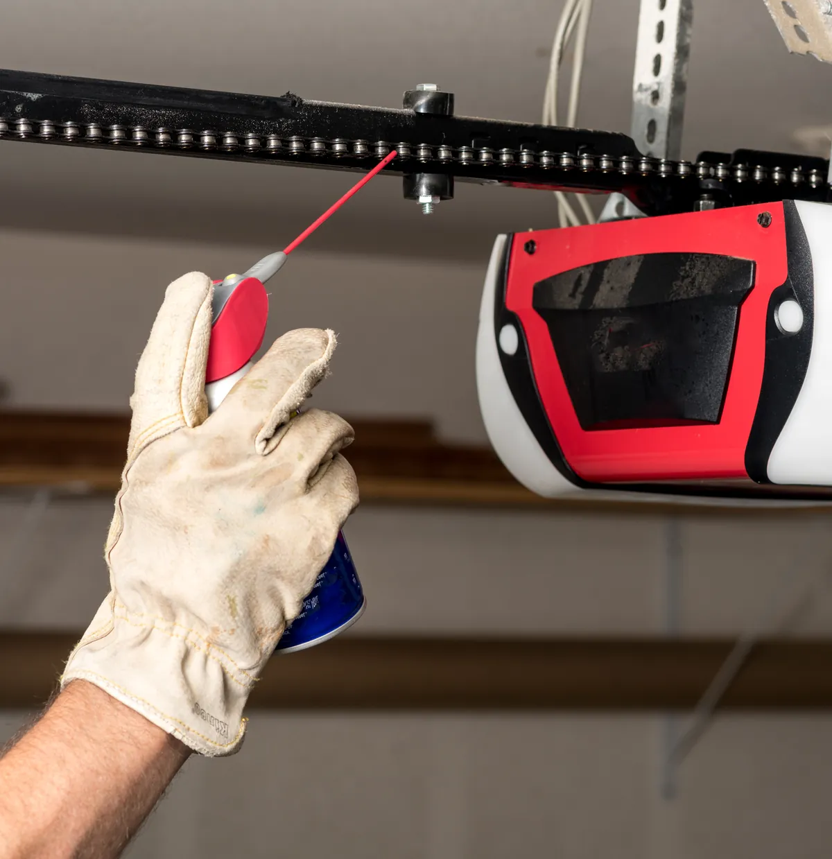 Gloved hand spraying lubricant onto a garage door opener's drive chain, beside the red and white opener unit