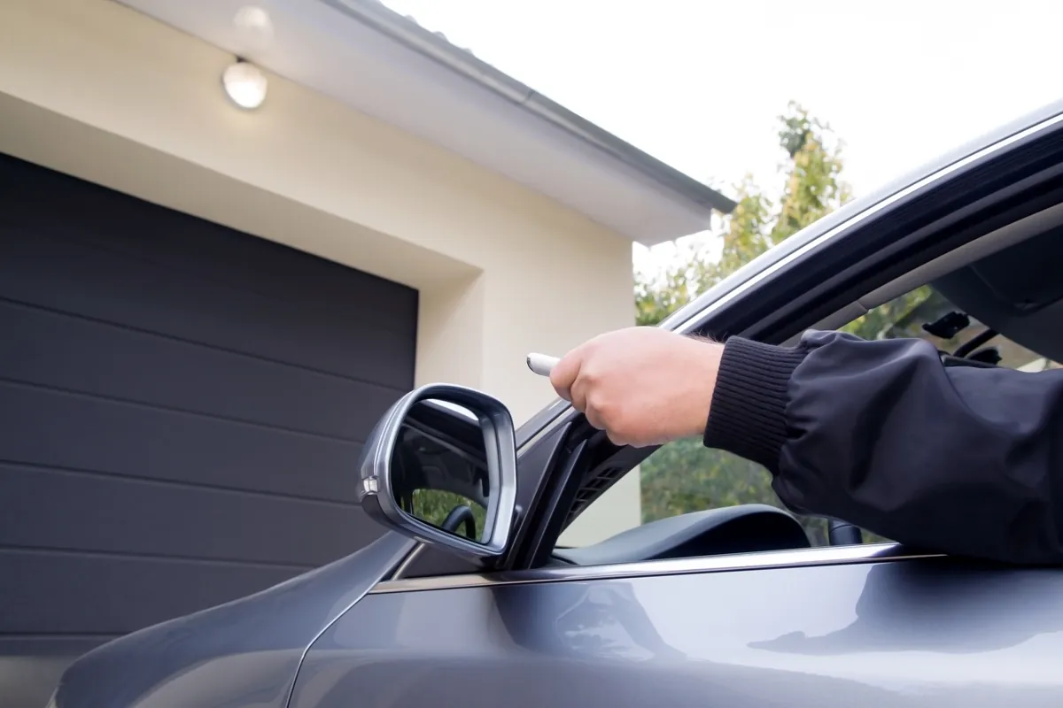 Driver pointing a remote from a car window toward a dark grey garage door