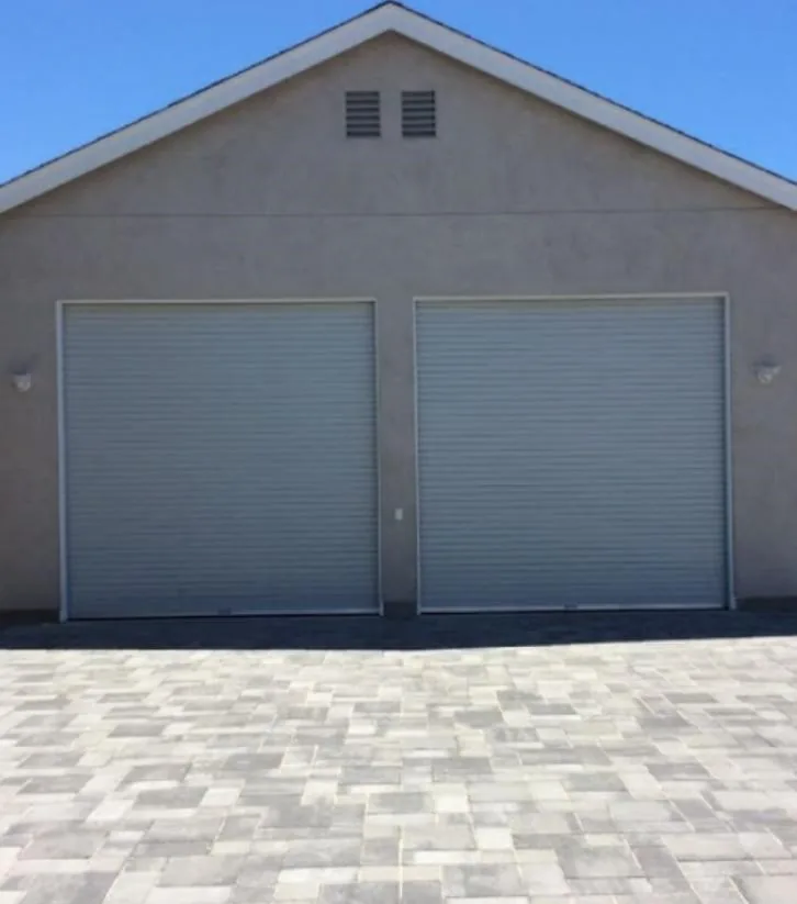 Two grey roll-up garage doors on a stucco garage above a paver driveway