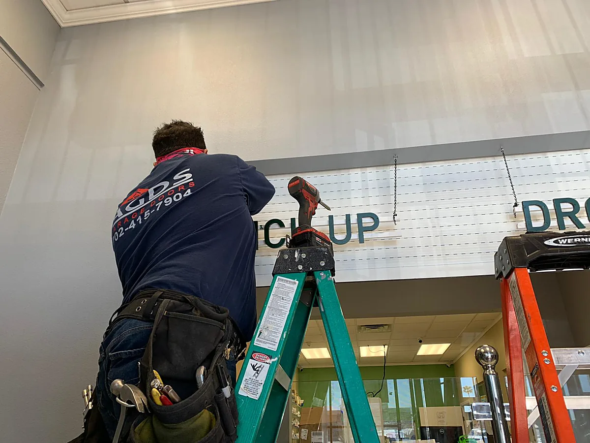 Technician with a tool belt working from a ladder at a pickup-counter opening, his shirt printed with 702-415-7904